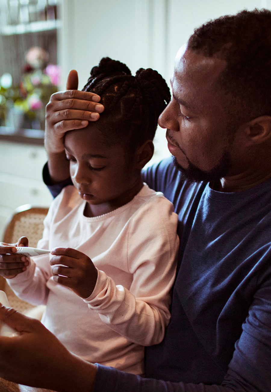 Little girl sitting on her father's lap while he checks her temperature with his hand.